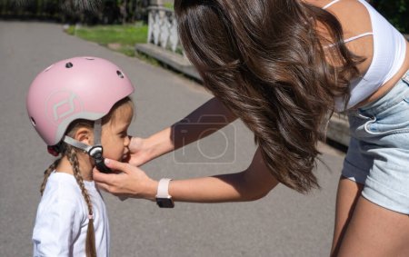 Photo for Mom putting on inline skates rollers protection helmet to daughter child in public park in summer. Family leisure outdoor sport activity game. - Royalty Free Image