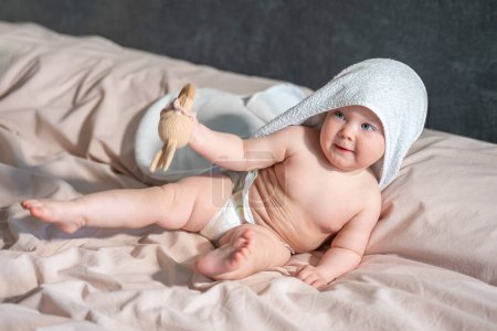Téléchargez les photos : Bébé dans la serviette de bain après les procédures d'hygiène, couché sur le lit parent dans la chambre. Maintenir peigne Hygiène et soins de la peau pour les enfants. - en image libre de droit