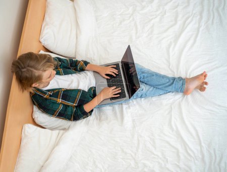 Photo for Teenage child boy using laptop. Child typing on the keyboard lying in bed. Play video game. Schoolboy studying doing homework. E-learning, remote educational classes top view - Royalty Free Image
