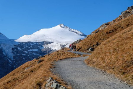 Téléchargez les photos : Sommet enneigé du Johannisberg, partiellement caché par une crête herbeuse brune avec un sentier de randonnée et quelques randonneurs - en image libre de droit