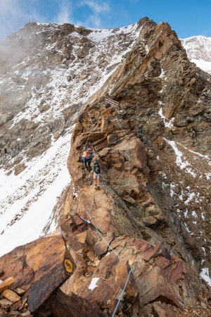 Téléchargez les photos : Deux randonneurs traversant une crête rocheuse exposée sécurisée par des cordes, neige visible sur les pentes voisines - en image libre de droit