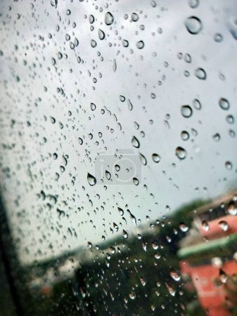 Foto de Grandes gotas de lluvia en el cristal de la ventana, durante la tormenta. Vista de las nubes nubladas y árboles y edificios verdes. La vista urbana de las gotas de lluvia cae sobre una ventana durante un día tormentoso. Lluvia en la ventana. Gotas de lluvia sobre vidrio. Gotas de lluvia en automoción - Imagen libre de derechos