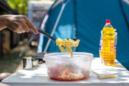 Foto de Anonymous male adding penne pasta into bowl while making pasta salad on campsite in summer - Imagen libre de derechos