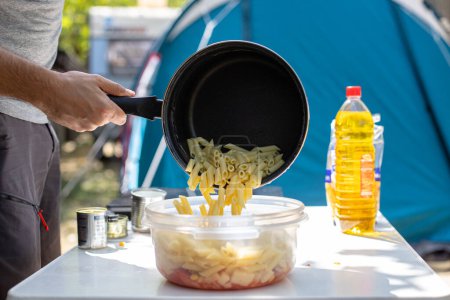 Foto de Unrecognizable man putting fresh penne pasta from saucepan into plastic bowl during lunch preparation on campsite - Imagen libre de derechos