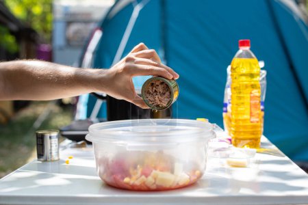 Foto de Anonymous male tourist adding canned tuna into salad in plastic bowl while preparing lunch on sunny day on campsite - Imagen libre de derechos