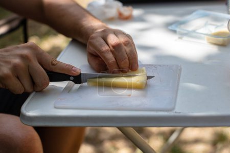 Foto de Side view of a seated male tourist cutting cheese on a white plastic cutting board preparing a picnic on a sunny day - Imagen libre de derechos