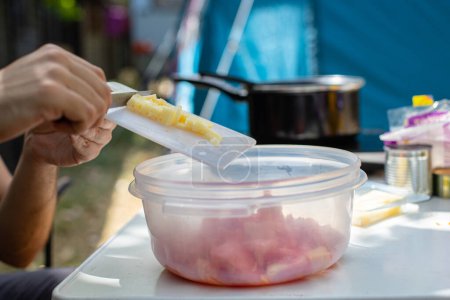 Foto de Unrecognizable man adding cut cheese into plastic bowl with tomatoes while sitting at table and cooking lunch on holidays on campsite - Imagen libre de derechos