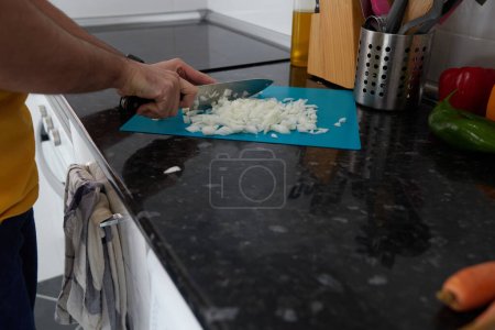 Foto de Anonymous person cutting an onion on a blue cutting board on a black countertop in a lighted kitchen - Imagen libre de derechos
