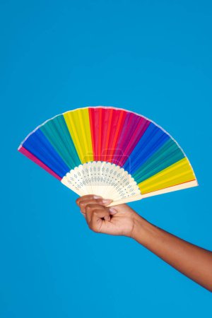 Foto de Crop unrecognizable female demonstrating bright colorful folding fan against blue background in studio - Imagen libre de derechos