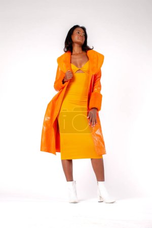 Foto de Full body self assured ethnic female model in trendy dress and coat touching hip and looking at camera against white background - Imagen libre de derechos