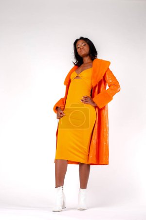 Foto de Stylish mixed race woman in yellow dress and orange coat with hand on waist touching neck and looking at camera against gray background - Imagen libre de derechos
