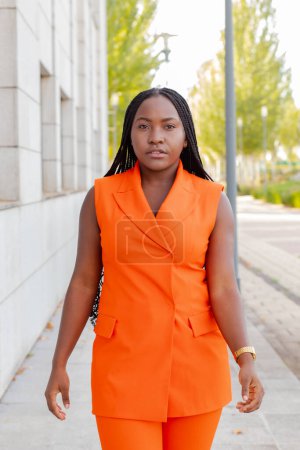Foto de Contemporary African American woman in trendy bright orange vest suit and with Afro braids walking on pavement looking at camera - Imagen libre de derechos