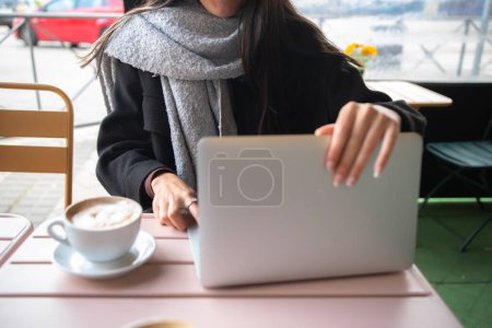 Foto de Unrecognizable woman in coat and scarf opening and turning on netbook near cup of coffee while sitting at table in street cafeteria - Imagen libre de derechos