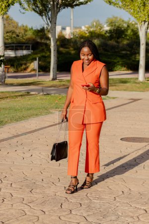 Foto de Modern African American lady in vivid orange suit checking mobile phone while standing on sunlit street - Imagen libre de derechos