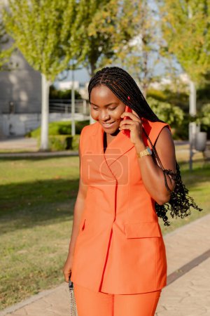 Foto de Content modern African American female in vivid orange vest suit and with Afro braids having call on mobile phone walking in sunlight - Imagen libre de derechos