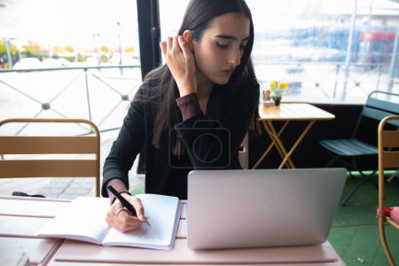 Foto de Young woman with long dark hair with notepad browsing data on laptop during work on remote project in street cafe - Imagen libre de derechos