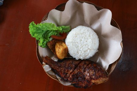 Photo for Grilled gourami or Gurame bakar with red barbeque sauce, rice, vegetables, tofu, and tempeh on a rattan plate. - Royalty Free Image