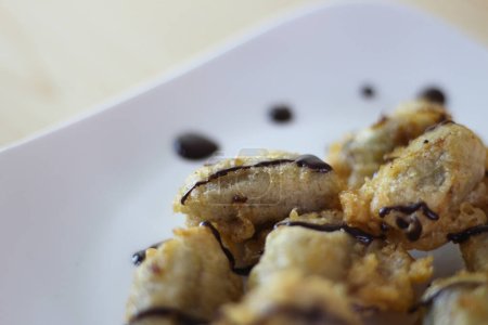 Closeup view of Pisang Goreng Keju or Fried Banana topped chocolate with on white plate on the wooden table.
