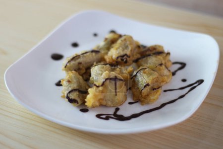 Pisang Goreng Keju ou banane frite surmonté de chocolat avec sur plaque blanche sur la table en bois.