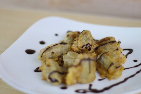 Pisang Goreng Keju or Fried Banana topped chocolate with on white plate on the wooden table.