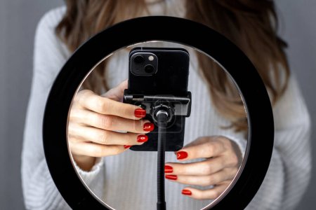 Téléchargez les photos : Woman girl young blogger vlogger talking with followers hand holding phone in middle of selfie light round lamp.blonde hair red nails female explaining moving hands from side to side or typing - en image libre de droit