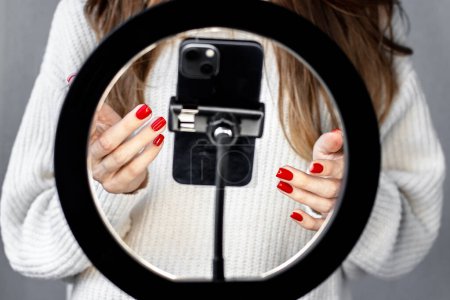 Foto de Woman girl young blogger vlogger talking with followers hand holding phone in middle of selfie light round lamp.blonde hair red nails female explaining moving hands from side to side or typing - Imagen libre de derechos