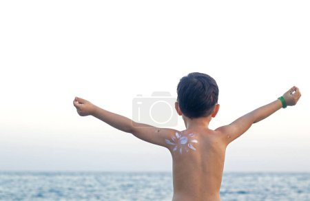 Photo for Kid boy preschooler with drawn sun shape from cream lotion on shoulder against sea ocean tan red skin hand up family vacation summer time.sun block protection white cream uv rays safe for skin care - Royalty Free Image