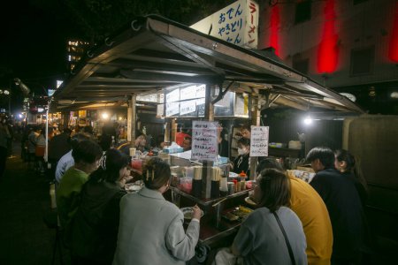 Foto de Fukuoka, Japón; 1 de octubre de 2023: Personas comiendo y bebiendo en un Yatai en la bahía de Hakata. Un Yatai es un pequeño puesto de comida móvil en Japón que típicamente vende ramen u otros alimentos.. - Imagen libre de derechos