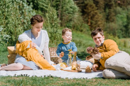 A young family with two children sits on a plaid and plays on a country lawn enjoys a picnic in sunny weather, copyspace, all dressed on yellow, blue and white clothes, time to family weekend concept