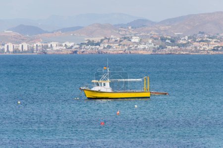 Photo for A small yellow boat with the Spanish flag sails in the Mediterranean Sea. In the background, mountains and a picturesque village can be seen. This peaceful image showcases the natural beauty of the area. - Royalty Free Image
