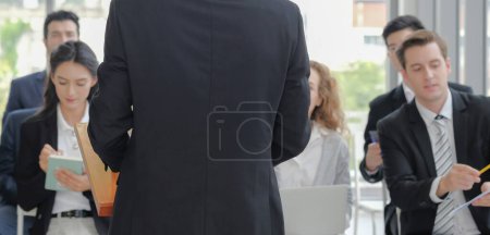 Photo for Back view of business male speaker in suit stand on podium and making speech, discussing while audience listening and taking notes in corporate seminar event. Selective focus. - Royalty Free Image