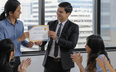 Photo for Young caucasian manager in suit giving mock up certificate to asian staff with happy smiling face while others applauding for congratulation in office meeting room. Selective focus. - Royalty Free Image