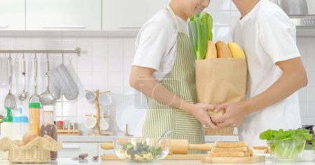 Photo for Young LGBT couple domestic life concept. Half portrait young LGBT male couple happy holding shopping bag with ingredients together during cooking in the kitchen. Selective focus and copy space. - Royalty Free Image