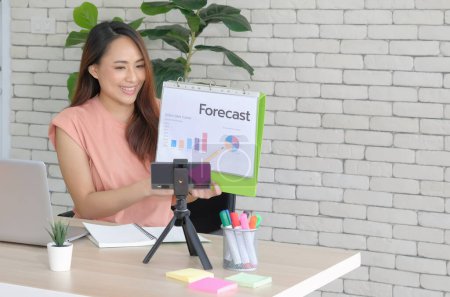 Téléchargez les photos : Jeune asiatique casual business femme confiant sourire lors de la présentation du rapport de prévisions de ventes sur smartphone en direct en streaming au bureau. - en image libre de droit
