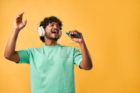 Portrait of handsome curly Indian man in turquoise t-shirt and singing with phone in wireless headphones enjoying music and gesturing emotionally.