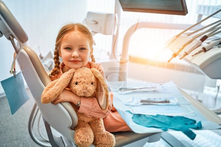 Photo of a little girl sitting in a dental chair, holding a toy rabbit and laughing. Dental care, medicine. Copy space.Sunlight.                               