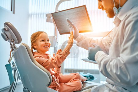 Ein kleines Mädchen sitzt auf einem Zahnarztstuhl, gibt dem Arzt eine hohe Fünf und lacht. Zahnpflege, Vertrauen und Patientenversorgung. Kinderzahnärzte.Sonnenlicht.                               