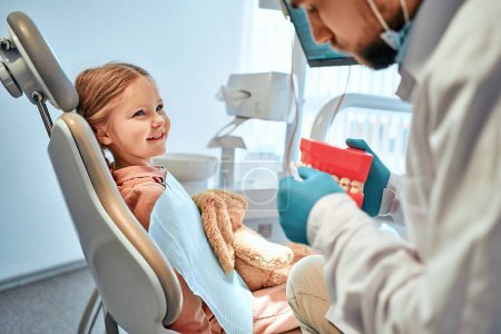 Photo for Children's dentistry. A little girl sits in a dental chair and looks at the doctor who shows her a model of the jaw and shows how to brush her teeth. - Royalty Free Image