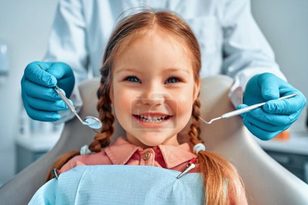 Photo for Cropped portrait of little girl with pigtails hair sitting in dental chair looking at camera and smiling. Behind, a doctor in gloves holds examination tools.Children's dentistry. - Royalty Free Image