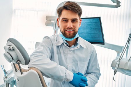Retrato de un médico dentista con frenos y una bata blanca, máscara y guantes sentados en el consultorio dental y mirando a la cámara y sonriendo.