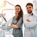 A team of doctors in a dental office standing back to back and looking at the camera and smiling. The work and leisure of doctors. In the background, a dental chair and equipment. Copy space.Sunlight.