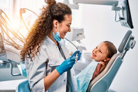 Téléchargez les photos : Une dentiste parle à une petite patiente, l'enfant montre ses dents et s'assoit sur le fauteuil dentaire.. - en image libre de droit