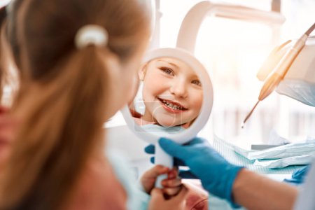 Une petite fille est patiente chez le dentiste. Un enfant tient un miroir, regarde ses dents et sourit..