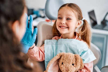 Au rendez-vous chez le médecin. Une photo émotionnelle franche d'un enfant assis dans une chaise dentaire, tenant un lapin jouet et donnant joyeusement un high-five à l'infirmière.