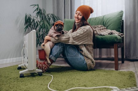 Foto de Una joven hembra vestida con calor y su perro se están calentando cerca de un radiador eléctrico. - Imagen libre de derechos