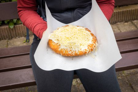 Photo for Woman holding a traditional langosh in her hands, sitting on a park bench, Langos, a deep fried pastry popular in Eastern Europe, served with toppings such as cheese and yogurt, Street food - Royalty Free Image