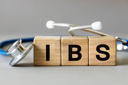 Medizinisches Stethoskop und Holzklötze mit der Aufschrift IBS, Abkürzung für Reizdarmsyndrom, eine chronische Erkrankung des Magen-Darm-Traktes funktioneller Natur, grauer Hintergrund