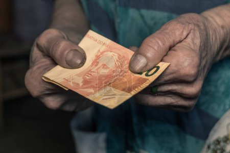 Underpaid job in Brazil, Concept, Old woman holding 20 reais banknote in her worn hand, Gloomy dark colors