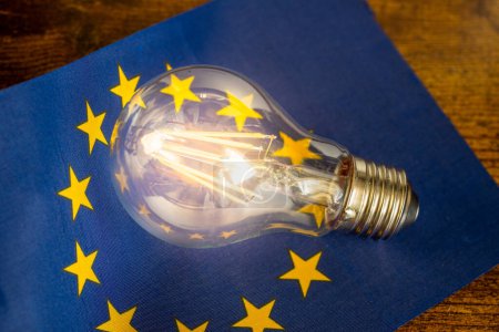 Eine glühende Glühbirne, die in der Mitte der Flagge der Europäischen Union liegt, Konzept, Energiepreise in EU-Ländern, Änderungen aufgrund der Energiepolitik