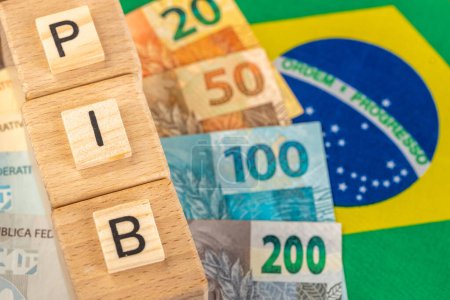 Das Wort PIB (Bruttoinlandsprodukt) steht auf Holzwürfeln mit brasilianischem Echtgeld in gelb-grüner Flagge Brasiliens. Brasilianisches Portugiesisch